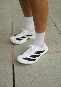 Persoană îmbrăcată cu pantofi sport albi cu dungi negre și șosete albe, stând pe trotuar gri din plăci.