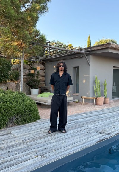 Camisa negra de manga corta con un bolsillo, pantalones sueltos negros y sandalias negras. Tiene el cabello rizado y gafas de sol; está de pie en una terraza de madera.