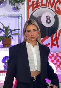 Crni skraćeni sako preko bijele košulje s kragnom, uparen s tamnim hlačama. Pozadina sadr�ži disko kugle, biljke i šarenu umjetnost.