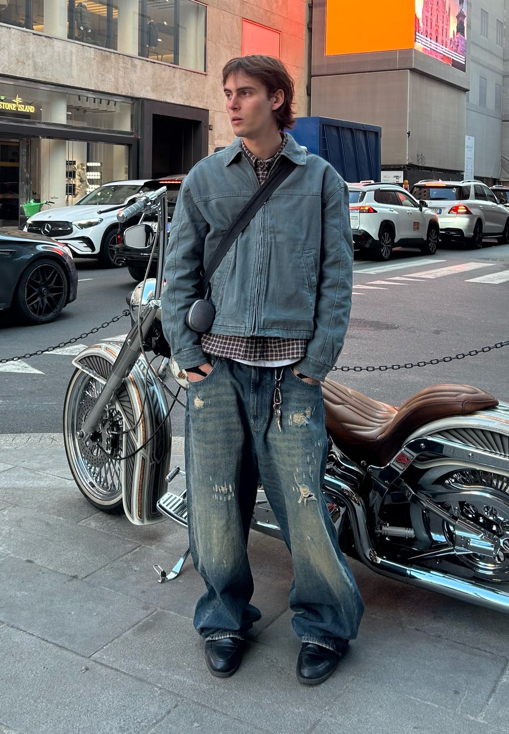Jaqueta de denim azul desbotada, camisa xadrez e calças largas desgastadas; em pé perto de uma motocicleta cromada numa rua da cidade.