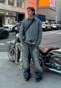 Jaqueta de denim azul desbotada, camisa xadrez e calças largas desgastadas; em pé perto de uma motocicleta cromada numa rua da cidade.