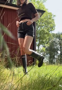Schwarze Gummiregenstiefel mit strukturiertem Grip, einem abgerundeten Design und einem Logo an der Seite. Getragen mit dunklen Shorts in einem bewachsenen Bereich.