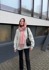 Túlméretezett világosszürke kötött pulóver, rózsaszín rojtos sál, fekete nadrág és egy kis fekete táska. Egy modern csempézett épület előtt áll.