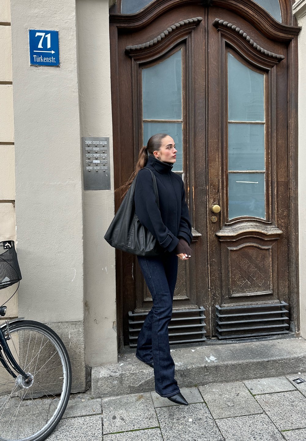 Čierna kapucňová bunda, tmavé džínsy a špicaté topánky. Stojí pri hnedých dvojitých dverách, viditeľné bicykel a interkomová panel. Ulicná značka na stene.
