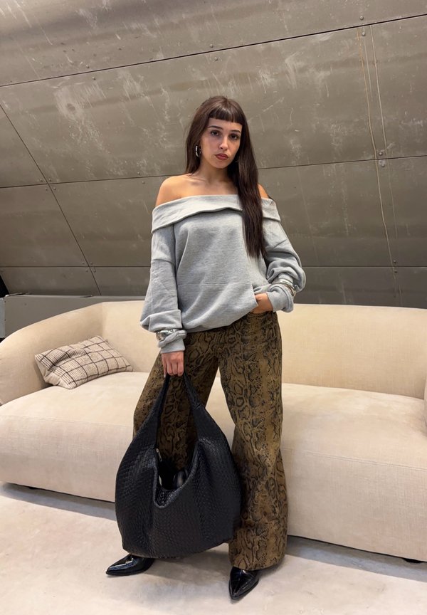 Mujer con cabello largo y castaño lleva un suéter gris con los hombros descubiertos y pantalones con estampado de serpiente, sostiene un bolso grande negro, está de pie frente a un sofá beige.