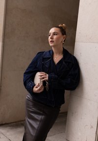 Jeansjakke med krave, lange ærmer og bælte i taljen, parret med en brun lædernederdel og en vævet beige clutch.