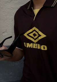 Hnědá polokošile s žlutým logem Umbro, s texturovaným vzorem a kontrastním žlutým límečkem. Černé sluneční brýle drženy v ruce.