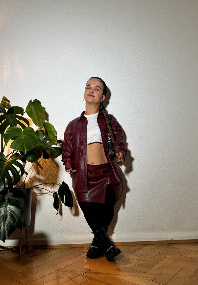 Μαρόν ψεύτικο δερμάτινο μπουφάν πάνω από ένα λευκό crop top, συνδυασμένο με μια ταιριαστή φούστα και μαύρες μπότες μέχρι το γόνατο, μπροστά σε έναν ουδέτερο τοίχο.