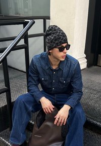 Camisa de ganga e calças em azul-escuro, gorro preto com padrão, óculos de sol e uma mala castanha. O padrão circular no gorro é notável.
