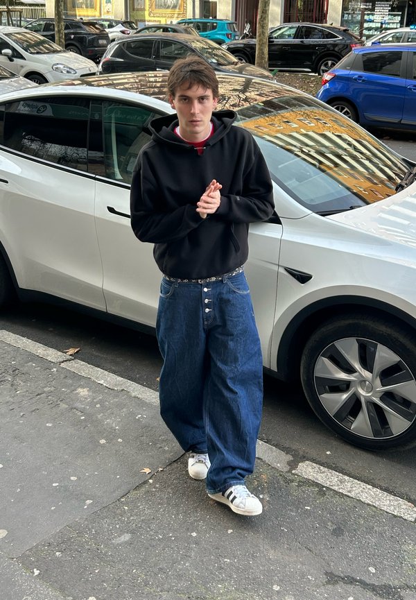 Jong persoon in een zwarte hoodie en wijde spijkerbroek staat op het trottoir naast een witte elektrische auto geparkeerd in een stadsstraat.