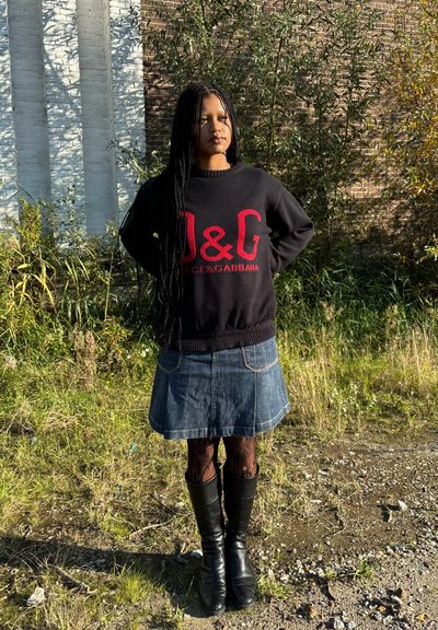 Suéter negro con marca roja, falda de mezclilla, botas altas negras, cabello largo trenzado, de pie en un área con hierba y follaje.