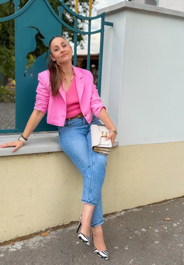Mujer con chaqueta rosa y vaqueros azules sentada en un muro bajo cerca de una puerta metálica azul con diseño de cabra, sosteniendo un bolso blanco.