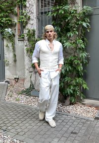 Persoană îmbrăcată într-un vest din in alb peste o cămașă albă și pantaloni lejeri albi, asortați cu adidași bej, pe fond de foliaj verde.
