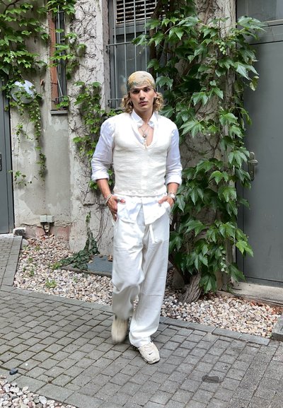 Persona vistiendo un chaleco blanco de lino sobre una camisa blanca y pantalones anchos blancos, combinados con zapatillas beige, con un fondo de follaje verde.