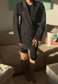 Personne portant une robe blazer noire, des leggings noirs, des bottes hautes marron clair et un petit sac scintillant, debout dans un salon éclairé par le soleil.