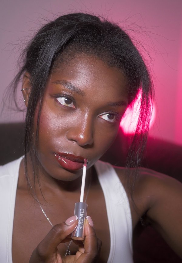 Une femme applique un gloss à lèvres Clinique à l’aide d’un applicateur, sur un fond éclairé en rose.