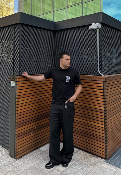 Camiseta negra con gráfico, pantalones negros holgados, zapatos brillantes, de pie junto a un panel de madera y una pared oscura texturizada. Ambiente urbano con reflejos.