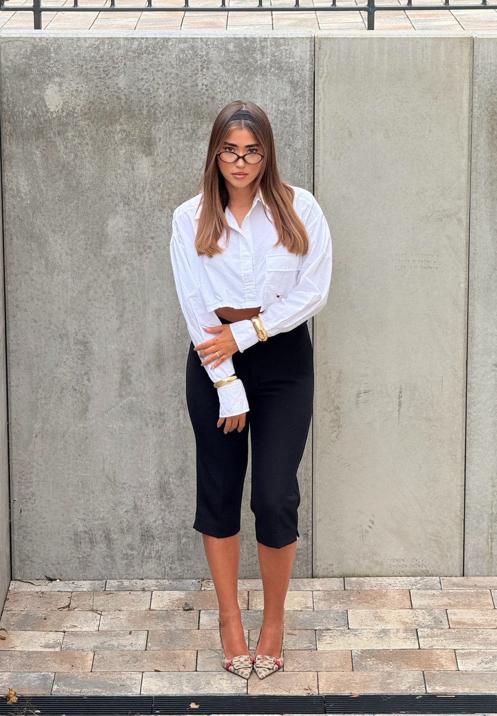 Camisa blanca corta, pantalones crop negros ajustados y tacones altos con estampado. Accesorios incluyen una pulsera dorada y gafas. Fondo neutro.