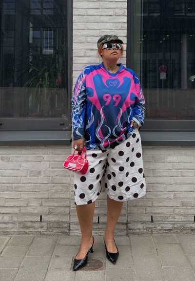 Persona que lleva una camiseta azul y rosa con estampado y número 99, pantalones cortos blancos con lunares negros, tacones negros, gafas de sol y sosteniendo un pequeño bolso rojo.