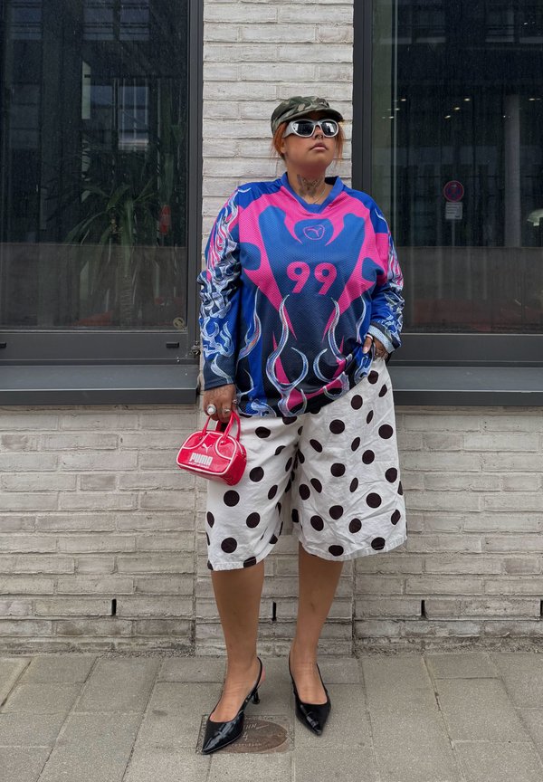 Persoană purtând un top albastru și roz cu model și numărul 99, pantaloni scurți albi cu buline negre, tocuri negre, ochelari de soare și ținând o geantă roșie mică.