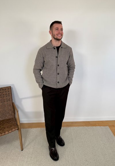 Hombre sonriente de pie con las manos en los bolsillos, vistiendo un cárdigan gris abotonado, pantalones negros y zapatos negros pulidos, junto a una silla de mimbre sobre una alfombra.
