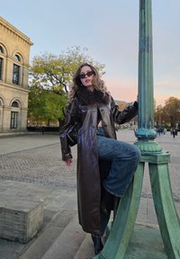 Dlhý hnedý kožený kabát s golierom z umelého kožušiny, vrstvený cez čierny top a modré široké nohavice, doplnený čiernymi slnečnými okuliarmi a členkovými topánkami.