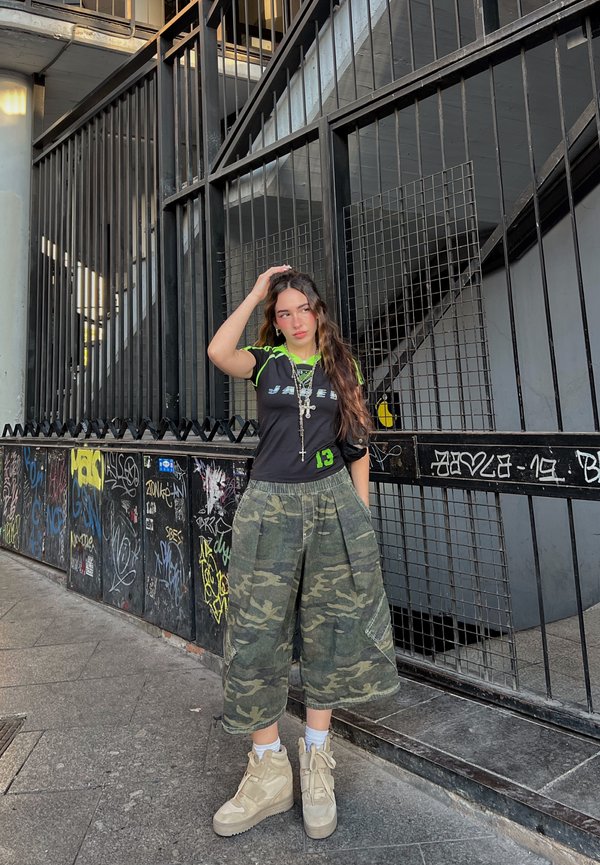 Cămașă neagră cu accente verzi și text grafic, asortată cu pantaloni scurți largi cu model camuflat și adidași bej înalți. Mediu texturat.