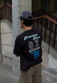 Osoba w ciemnej czapce z daszkiem i czarnej koszulce "Status Quo '91 Rock Till You Drop Tour" stoi obok kamiennych schodów i balustrady.