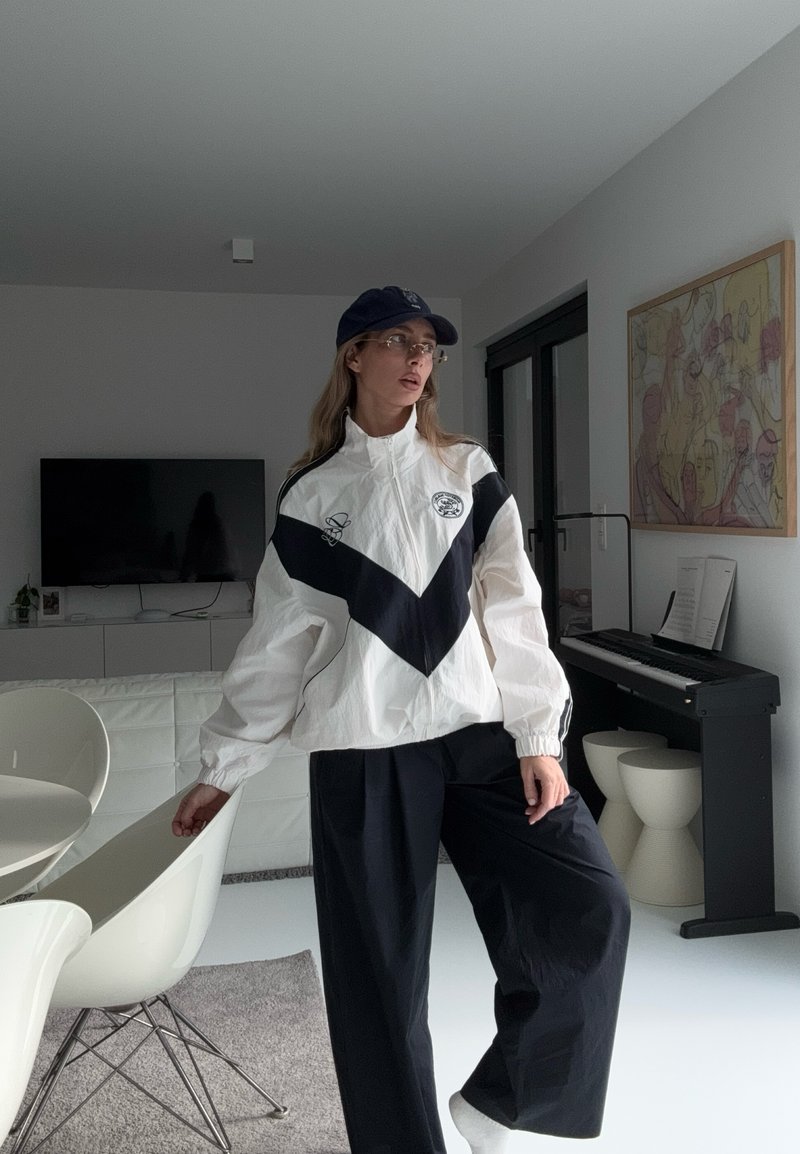 Γυναίκα με μαύρο καπέλο, γυαλιά, λευκό και μαύρο μπουφάν και μαύρο φαρδύ παντελόνι στέκεται σε μοντέρνο σαλόνι με πιάνο, τηλεόραση και αφηρημένη τέχνη τοίχου.