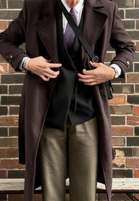 Καστανό κασκόλ από μάλλινη ύφανση φορεμένο πάνω από ένα μαύρο γιλέκο, γκρι παντελόνι με σχέδιο και καρό γραβάτα. Αξεσουάρ περιλαμβάνουν ένα ρολόι και μια τσάντα.