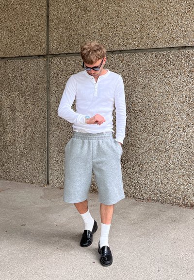 Camisa henley de manga larga blanca, pantalones cortos de sudadera grises con bolsillos, mocasines negros y calcetines blancos, contra un fondo de pared gris texturizada.