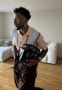 Homme portant un polo en tricot rayé et un pantalon marron, tenant une sacoche en cuir noir, debout dans un salon avec un canapé d'angle blanc et un sol en bois.