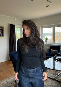 Kobieta z długimi ciemnymi włosami, ubrana w czarną bluzkę i niebieskie dżinsy, stoi w salonie z niebieskimi fotelami i oknem ukazującym jesienne drzewa.