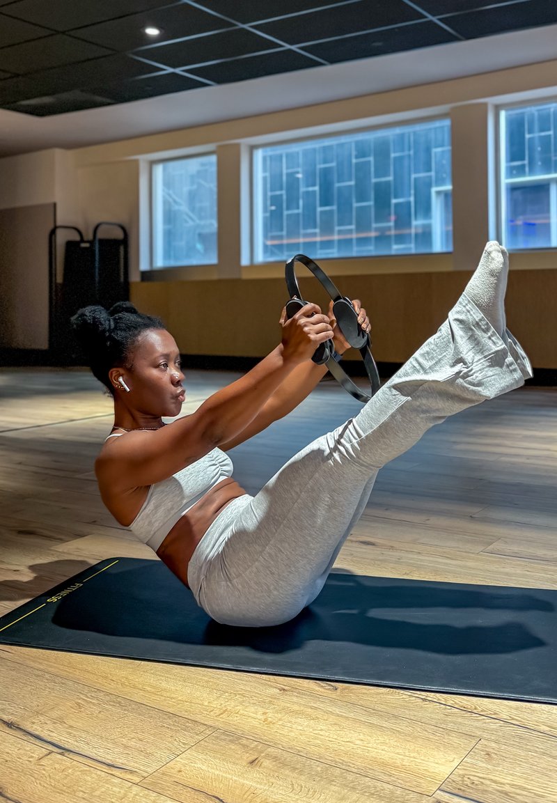 Femme en tenue de sport grise effectuant un exercice avec un cercle de Pilates sur un tapis dans un studio lumineux avec de grandes fenêtres et un sol en bois.