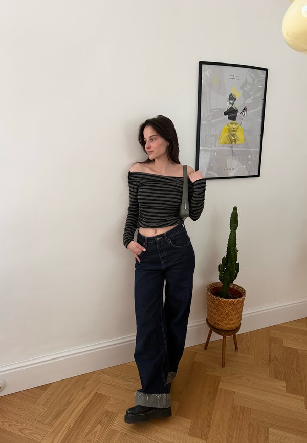 Νεαρή γυναίκα με ριγέ τοπ έξω από τους ώμους, σκούρα φαρδιά τζιν και μαύρες μπότες στέκεται σε ένα δωμάτιο με ξύλινο πάτωμα, κάκτο σε καλάθι και πλαίσιο με έργο τέχνης.