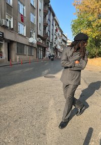 Sivasti prugasta obleka sproščenega kroja, črna bereta in črne čevlje. Urbano ulicno okolje z zgradbami in drevesi v ozadju.