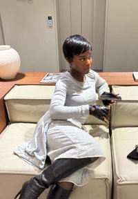 Robe grise à manches longues avec une jupe évasée, bottes noires montantes jusqu'aux genoux et gants sans doigts. Assise sur un canapé crème, tenant un téléphone.