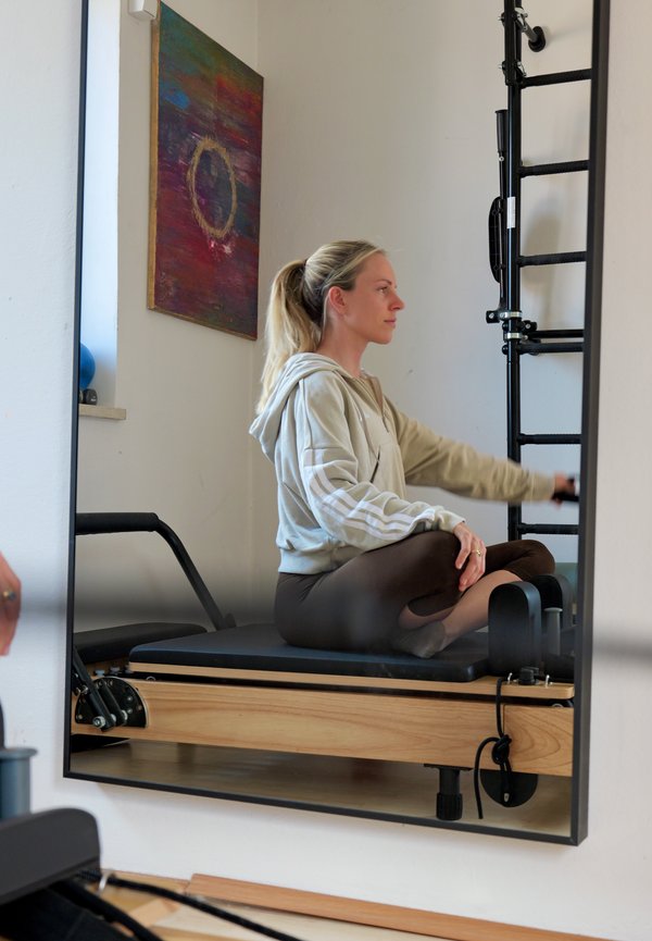Kobieta z kucykiem w bluzie z kapturem i legginsach siedzi po turecku na maszynie Pilates reformer, ciągnąc drążek oporowy w jasnym pomieszczeniu do ćwiczeń.