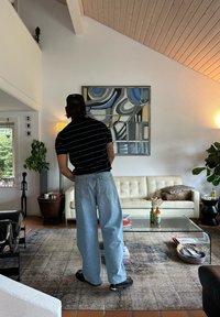 Persona con camisa de rayas negras y gorra de pie en una sala de estar moderna con sofá beige, mesa de café de vidrio, plantas y arte abstracto en la pared.