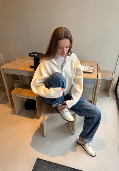 Suéter blanco acanalado con cremallera sobre una blusa clara, jeans de pierna ancha azules y zapatos tipo slip-on crema con acentos dorados. Fondo de muebles de madera.