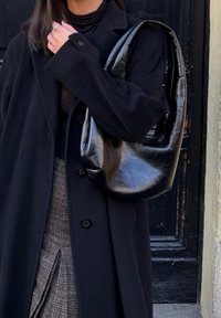 Manteau noir surdimensionné avec boutons, associé à une jupe à carreaux texturée. Un sac hobo noir brillant pend de l'épaule, mettant en valeur un matériau lisse.