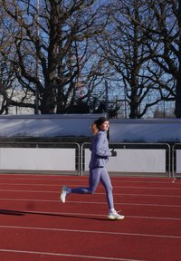 Tenue de sport bleu clair comprenant une veste zippée et des leggings. Chaussures de course blanches et jaunes. En train de courir sur une piste rouge sous un ciel dégagé.