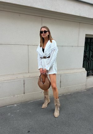 Prenda blanca con flecos y cinturón, bolso marrón y botas de cowboy beige con detalles de flecos. Fondo con una pared texturizada y pavimento.