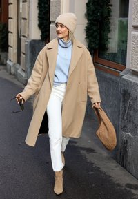 Manteau en laine beige, haut à fermeture éclair bleu clair, jean blanc et bottines beige. Tient un sac marron tressé et des lunettes de soleil. Cadre urbain avec végétation.