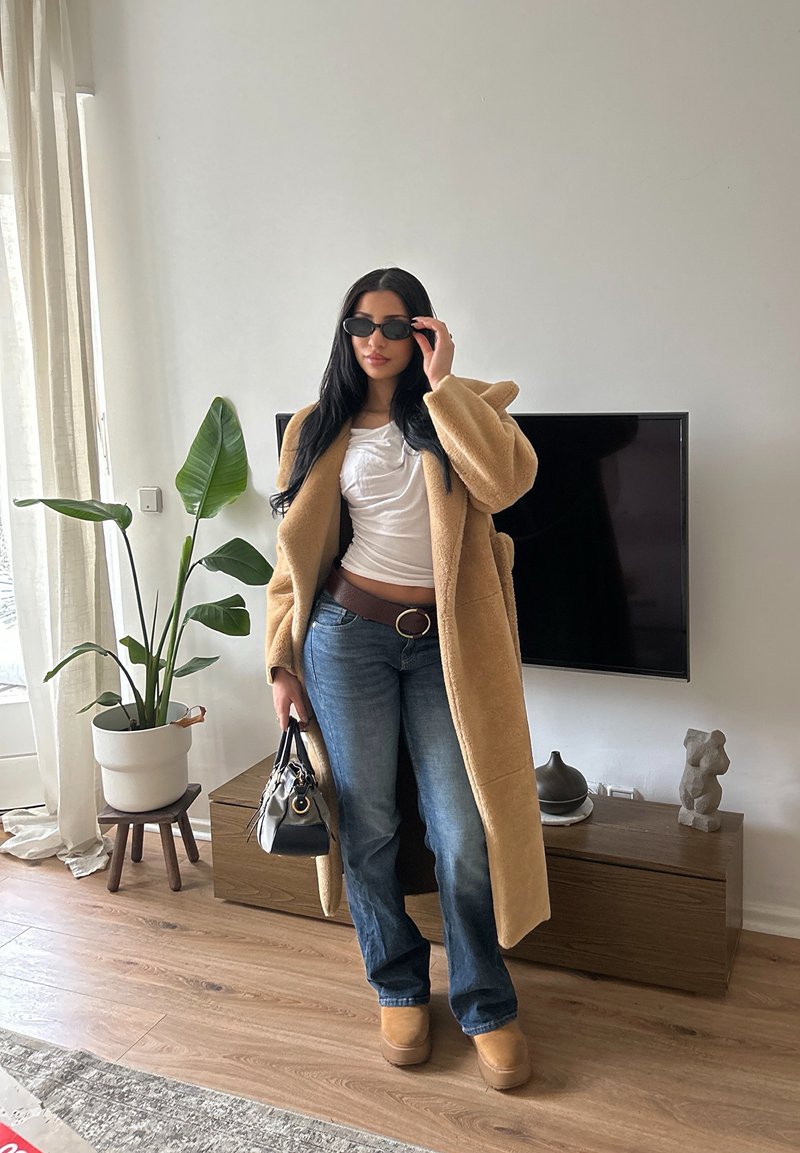 Femme portant des lunettes de soleil, un long manteau beige, un haut blanc, un jean bleu et des chaussures compensées, tenant un sac à main dans un salon moderne.