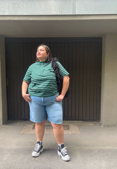 Camiseta a rayas verde y blanca, de corte holgado, combinada con pantalones cortos de mezclilla azul claro. Chaleco de piel negra caído sobre un hombro, zapatillas blancas con plataforma.