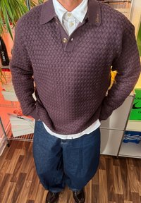 Bordowy sweter o fakturze dzianiny z kołnierzykiem i trzema guzikami, noszony na białej koszuli i zestawiony z niebieskimi, dużymi jeansami.