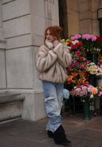 Palton din blană artificială de culoare bej deschis, design supradimensionat cu guler înalt; asortat cu blugi albaștri uzați și cizme negre cu franjuri până la gleznă, în mijlocul tarabelor cu flori.