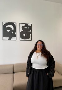Vrouw met lang krullend haar, die een bril draagt, een witte top, een zwarte jas en een rok, staat glimlachend naast een beige bank en twee abstracte zwart-witte kunstwerken.