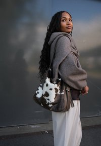 Vrouw in een bruine hoodie en lichte broek, met een grote schoudertas in een bruin-wit koeienhuidpatroon, staand tegen een grijze muur.